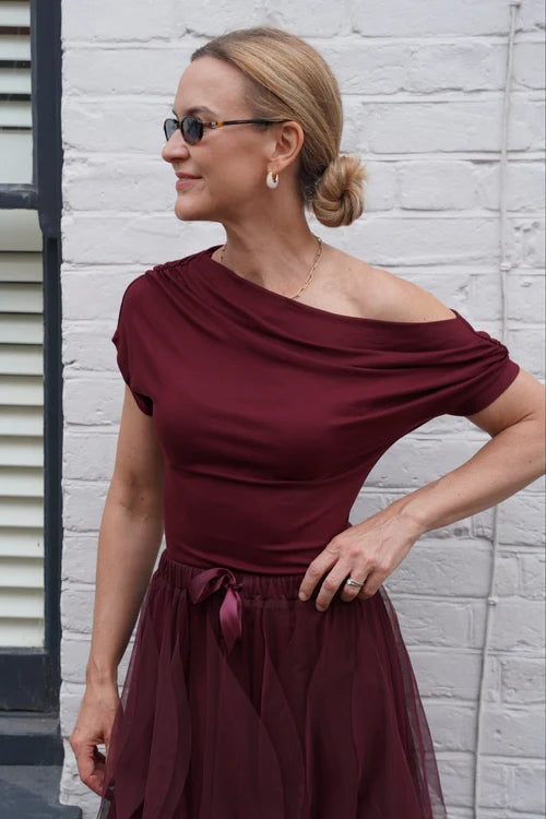 A woman wearing a burgundy off-the-shoulder dress with a sheer tulle skirt, standing against a white brick wall next to a window with shutters.