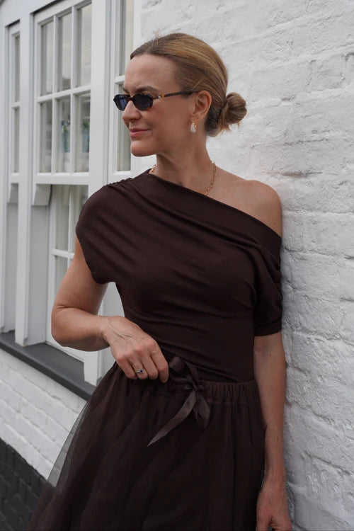 A woman wearing a dark brown off-the-shoulder midi dress with a sheer tulle skirt and a matching waist tie, posing against a white brick wall.