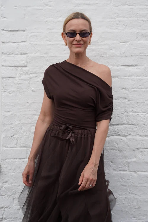A woman wearing a dark brown one-shoulder tulle dress and sunglasses, standing against a white painted brick wall.