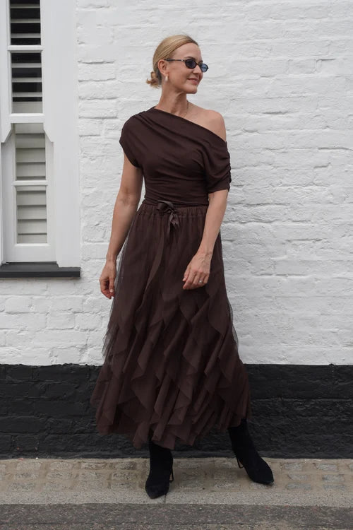 A woman wearing a dark brown one-shoulder midi dress with a ruffled tulle skirt, standing against a white brick wall.