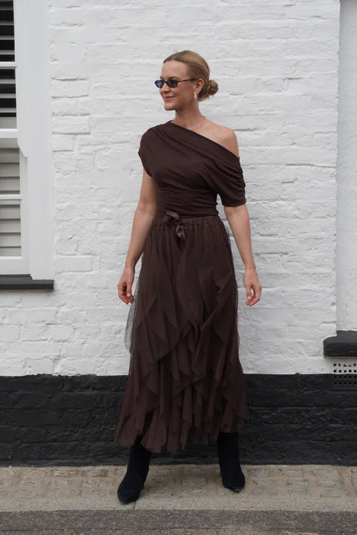 A woman wearing a dark brown one-shoulder midi dress with a ruffled tiered skirt, standing outdoors against a white brick wall.