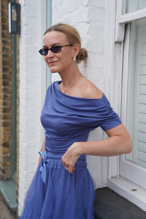 A woman wearing a blue off-the-shoulder midi dress and black sunglasses, posing outdoors against a white brick wall.