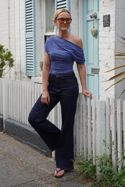 A woman wearing a blue off-the-shoulder top and dark navy wide-leg pants stands outdoors in front of a white brick house with a light blue door and shutters.