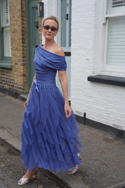 A woman wearing a blue off-the-shoulder tiered maxi dress and silver sandals walking on a street in front of a white brick house.