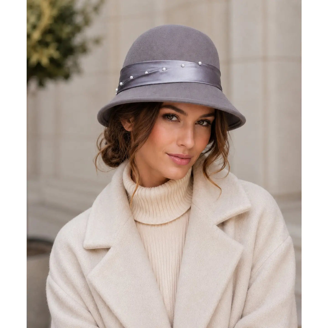 A woman wearing a gray felt cloche hat with a satin band and small pearl-like embellishments, paired with a cream-colored coat and turtleneck sweater, set against a soft beige stone wall background.