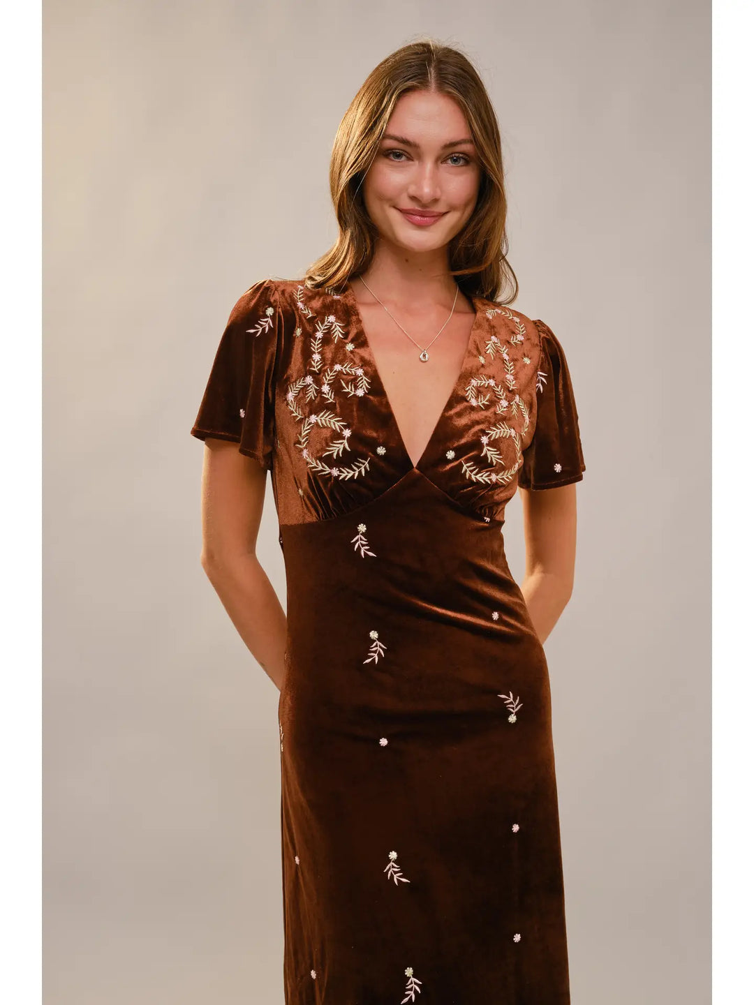 Woman wearing a brown velvet dress with floral embroidery on a plain background