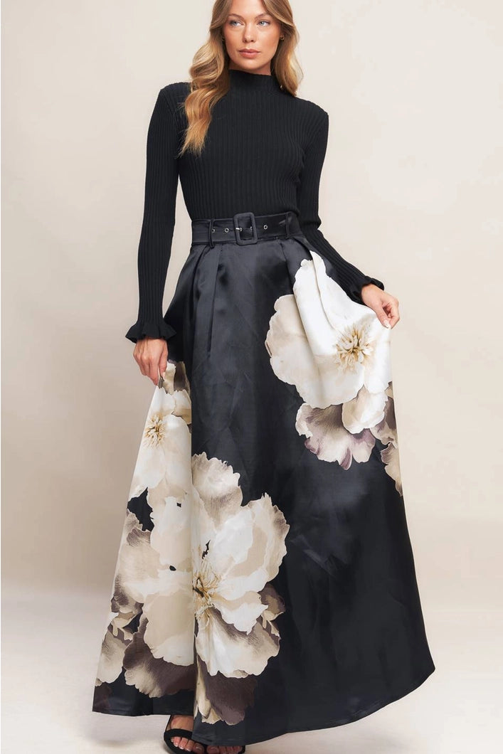 Woman wearing a black top and long black skirt with large white floral patterns on a plain background