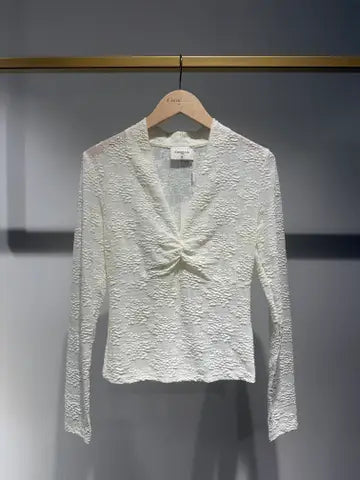 An off-white textured lace long-sleeve blouse hanging on a wooden hanger against a gray wall.