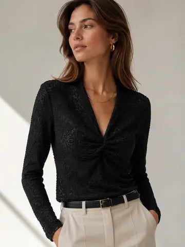 A woman wearing a black long-sleeve lace top with a twisted front detail, paired with cream trousers and a black belt, photographed against a neutral light gray background.