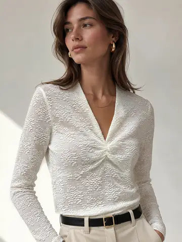 A woman wearing a fitted white textured long-sleeve blouse with a twisted V-neck detail, paired with beige trousers and a black belt, photographed against a light neutral studio background.