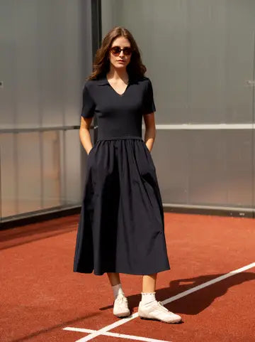 A woman stands on a red clay tennis court wearing a dark navy midi dress with short sleeves, a V-neckline, and a gathered waist, paired with white sneakers and sunglasses.