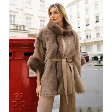 A woman wearing a brown fur wrap coat with a matching belt, standing next to a red post box on a city street with white buildings in the background.