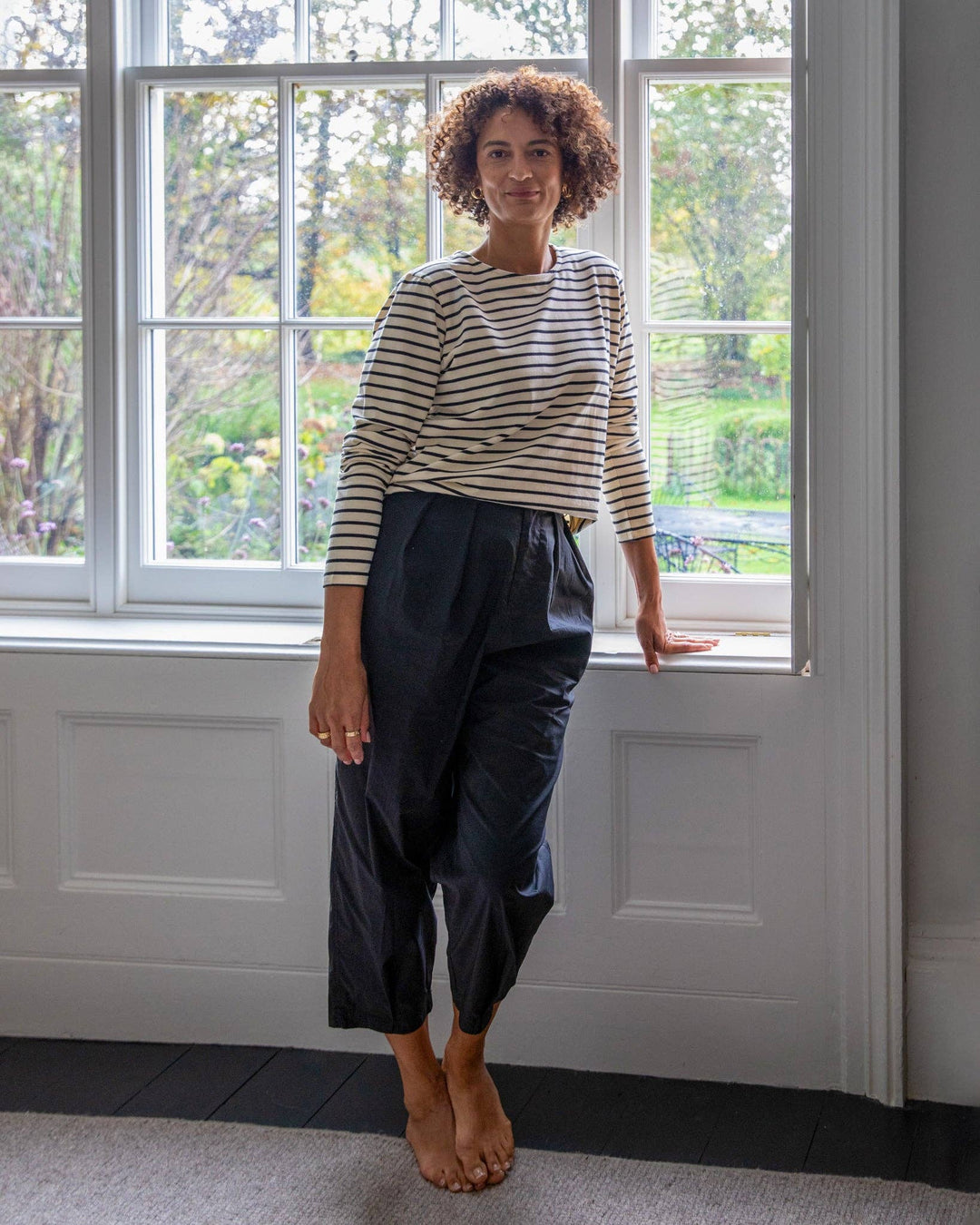 Woman wearing a striped sweater and dark pants standing in a room with large windows.