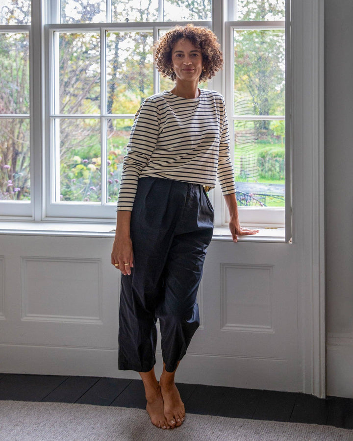 Woman wearing a striped sweater and dark pants standing in a room with large windows.