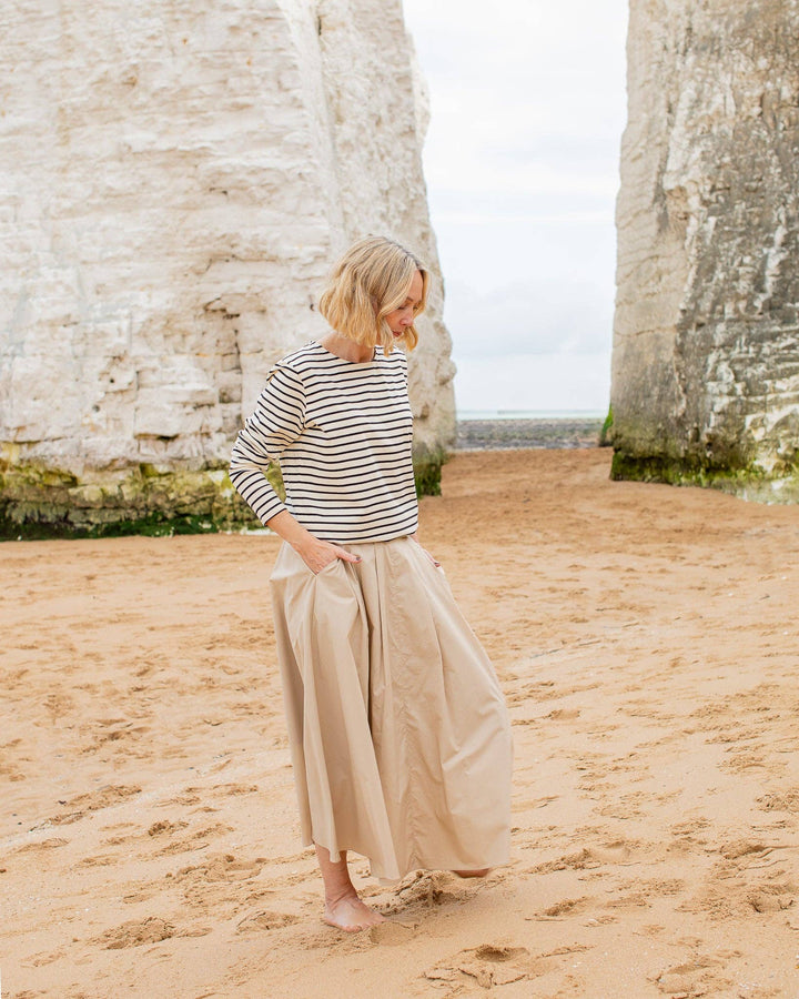 Woman in a striped shirt and beige pants standing on a sandy beach with rock formations in the background.