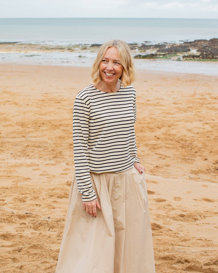 Woman standing on a beach wearing a striped sweater and beige skirt.
