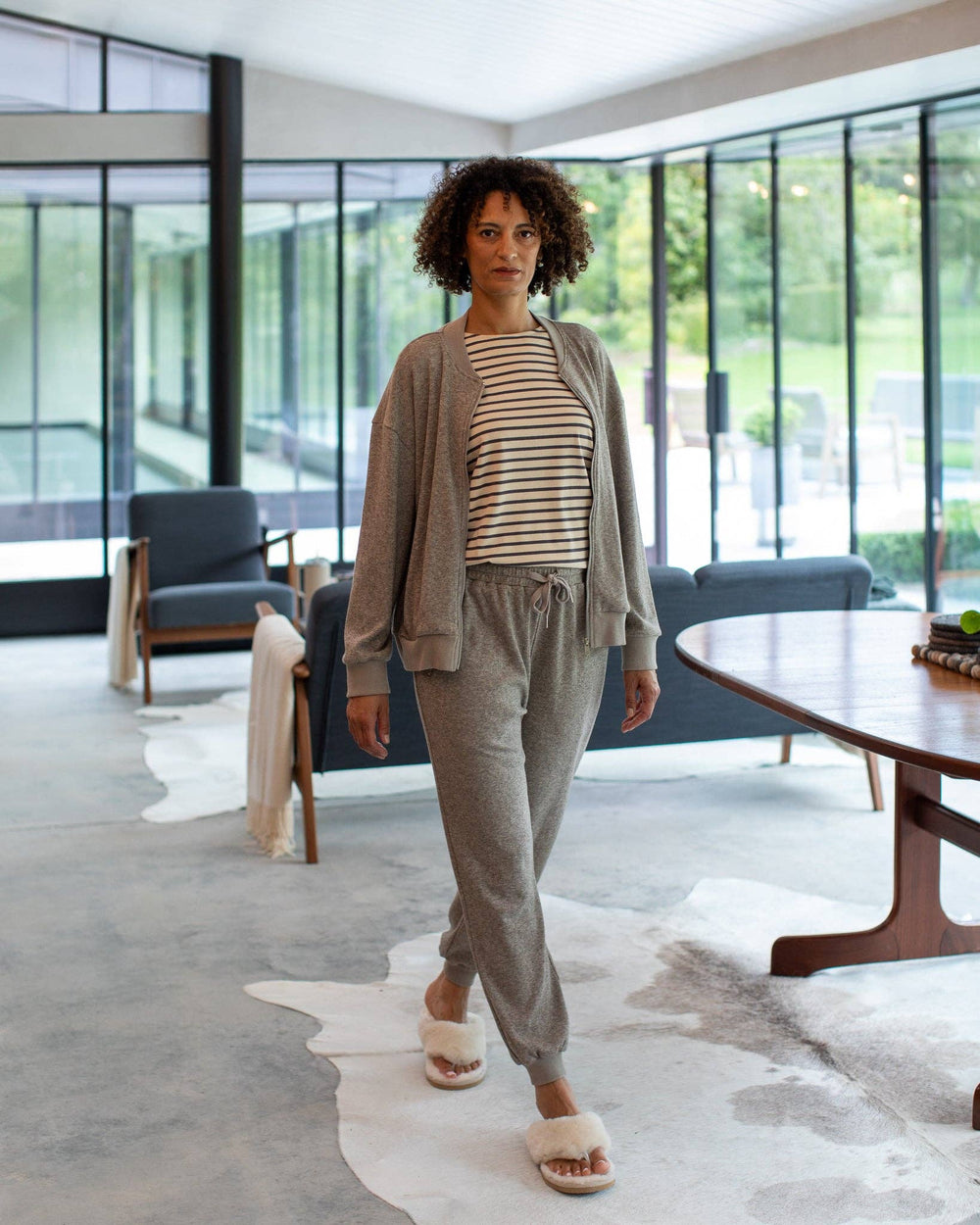Woman in a gray outfit standing in a modern living room with large windows.