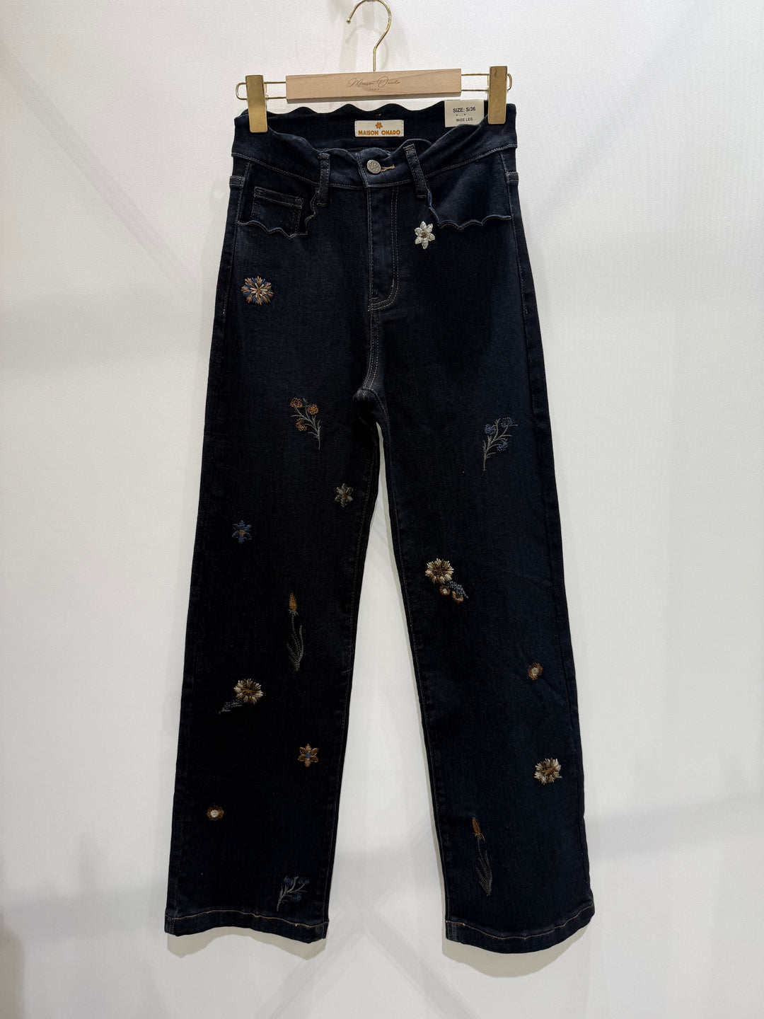 Dark indigo denim jeans with scattered embroidered floral details, hanging from a wooden hanger against a plain white wall.