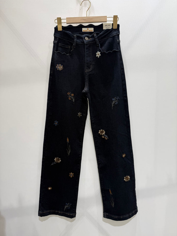 Dark indigo denim jeans with scattered embroidered floral details, hanging from a wooden hanger against a plain white wall.
