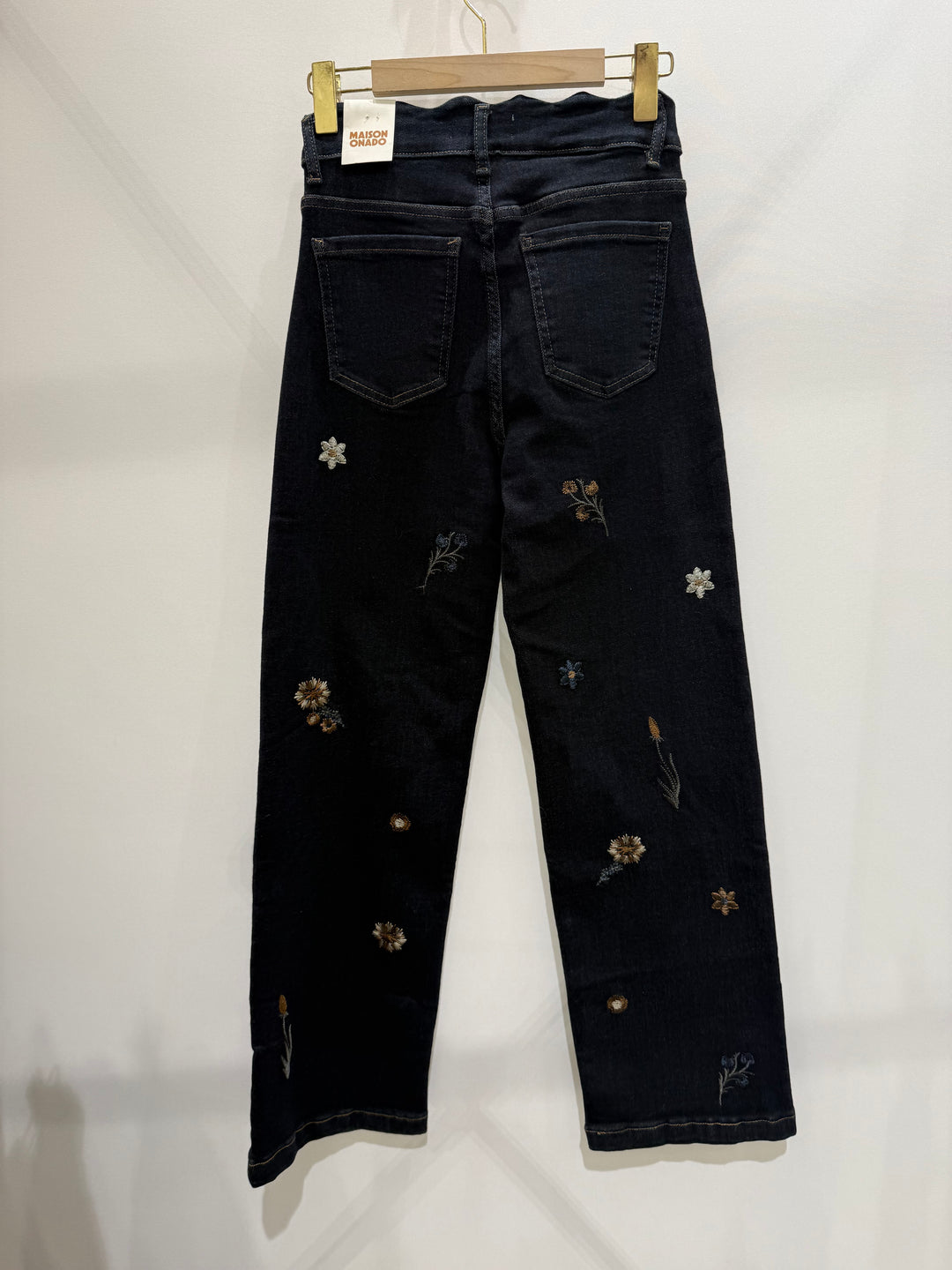 Dark wash denim jeans with embroidered wildflower motifs, hanging on a wooden hanger against a white wall.