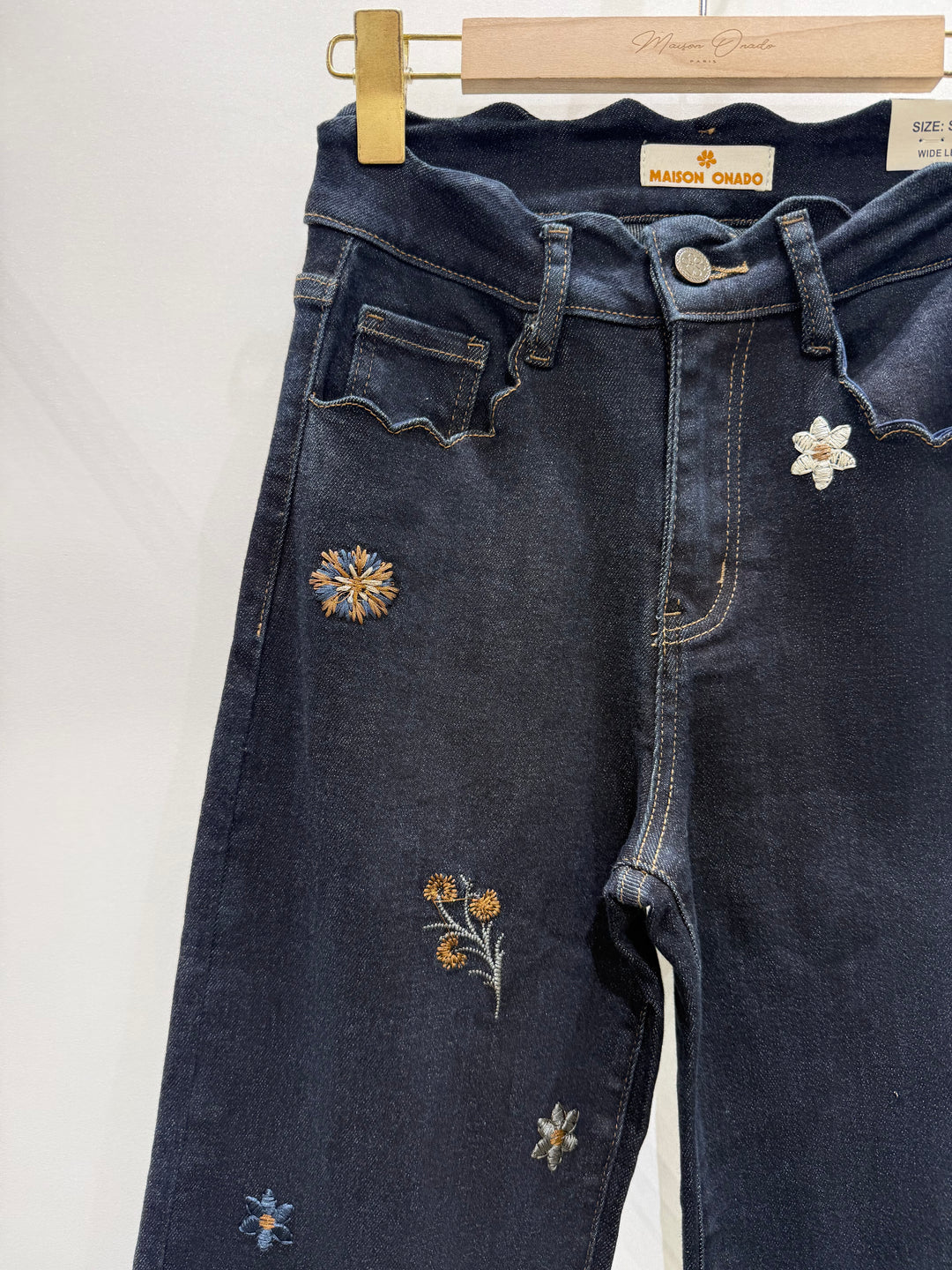 Dark blue denim jeans with embroidered floral details, hanging on a wooden hanger with a gold clip against a plain white background.