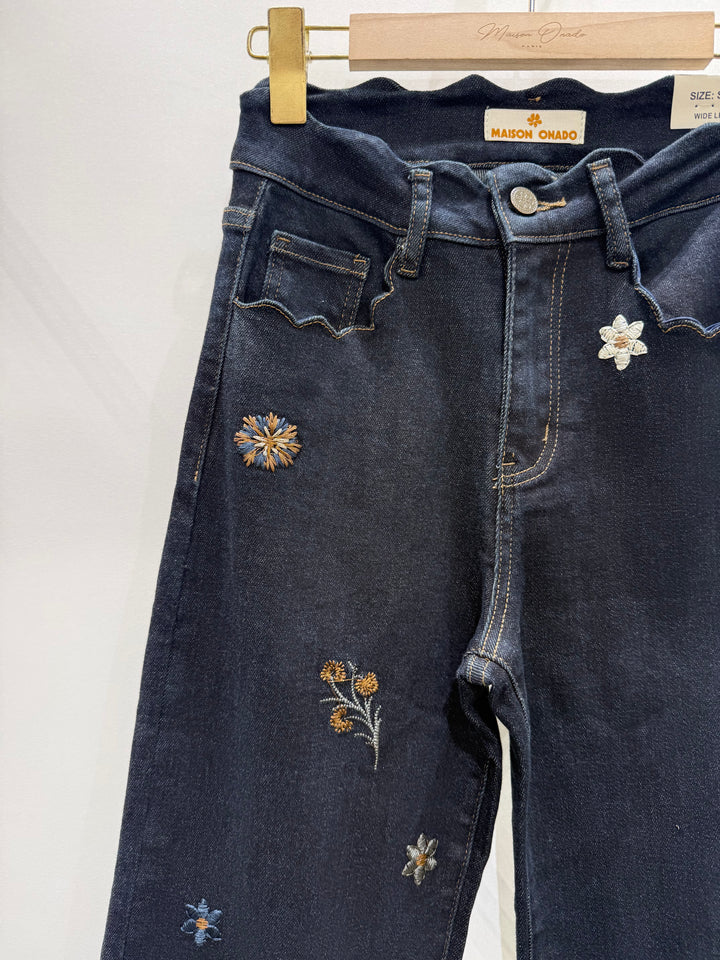 Dark blue denim jeans with embroidered floral details, hanging on a wooden hanger with a gold clip against a plain white background.