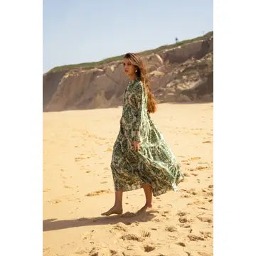 A woman wearing a green and white patterned maxi dress walks barefoot on a sandy beach with cliffs in the background.