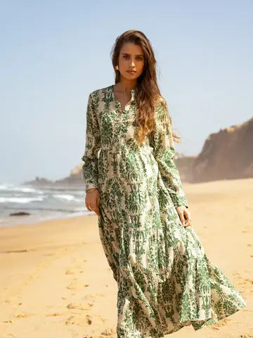A woman wearing a long-sleeved maxi dress with a green and beige botanical pattern walks along a sandy beach with the ocean and cliffs in the background.