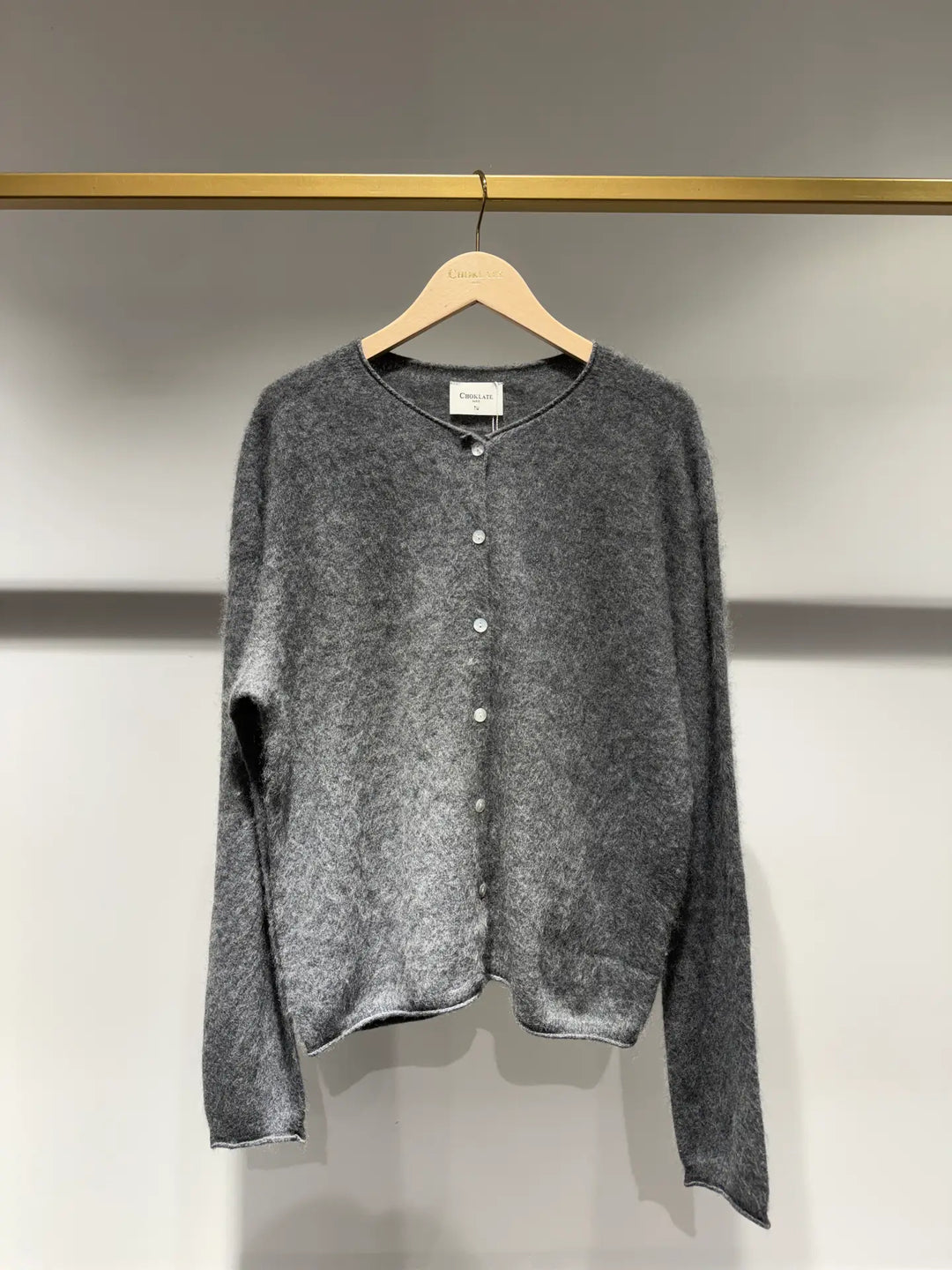 A gray fuzzy knit cardigan sweater with a button-up front, hanging on a wooden hanger from a gold clothing rack against a light gray wall.