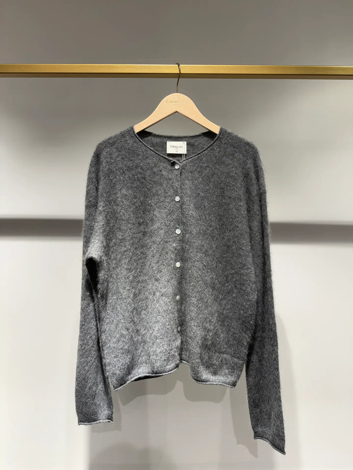 A gray fuzzy knit cardigan sweater with a button-up front, hanging on a wooden hanger from a gold clothing rack against a light gray wall.