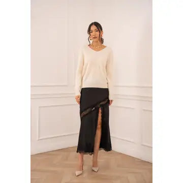 A woman modeling a cream-colored long-sleeve knit sweater paired with a black asymmetrical slit skirt featuring sheer lace trim, standing in a bright room with white paneled walls and light wood flooring.