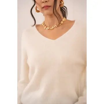 A close-up of a person wearing a chunky gold disc-link chain necklace and matching gold hoop earrings, paired with a soft cream-colored V-neck sweater.