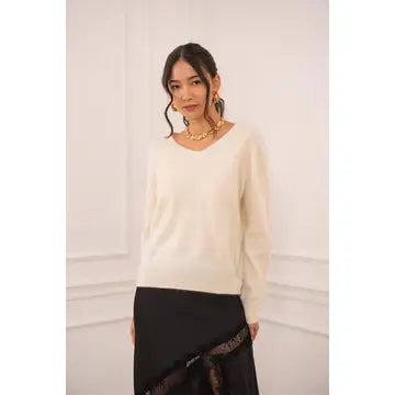 A woman wearing a cream-colored long-sleeve knit sweater and a black skirt, standing against a white paneled wall.