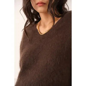A close-up of a person wearing a fuzzy brown V-neck sweater, with dark wavy hair and a gold chain necklace visible against a plain light background.
