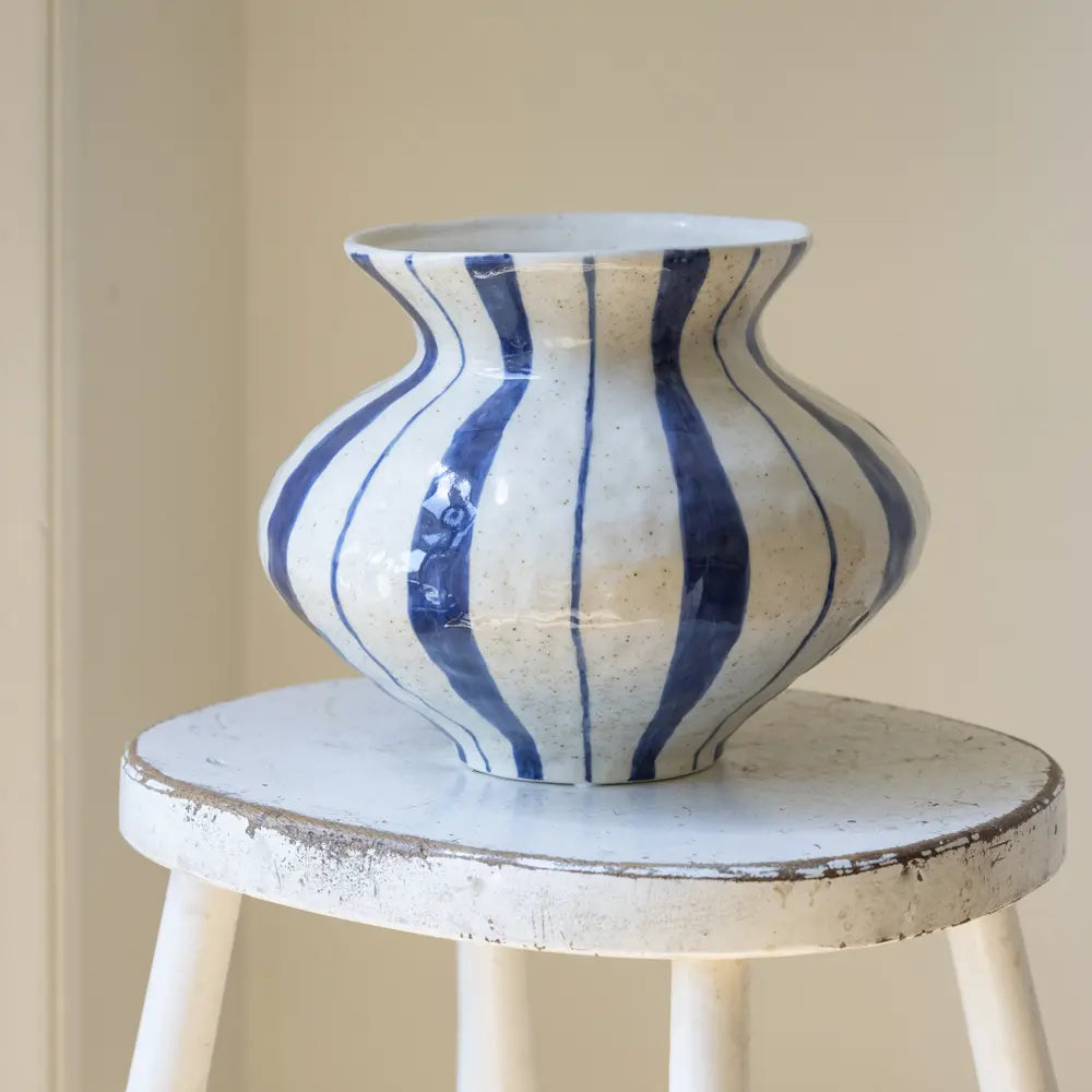 A ceramic vase with a bulbous shape, featuring vertical blue stripes on a speckled off-white glaze, displayed on a distressed white wooden stool against a neutral beige wall.
