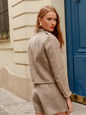 A woman wearing a beige tweed jacket and matching shorts set, photographed outdoors against a cream wall and a blue door.