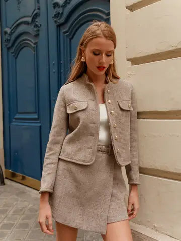 A woman models a light beige tweed jacket and matching mini skirt set with gold-tone buttons, styled over a white top, standing in front of a blue door and a cream-colored wall.