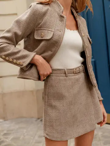 A woman models a matching beige tweed jacket and mini skirt set with a white ribbed tank top, photographed outdoors against a stone wall and blue door background.