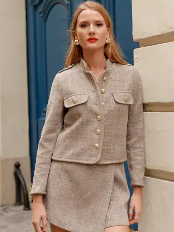 A woman models a beige tweed military-style jacket and matching skirt set with gold buttons, standing outdoors against a blue door and beige stone wall.