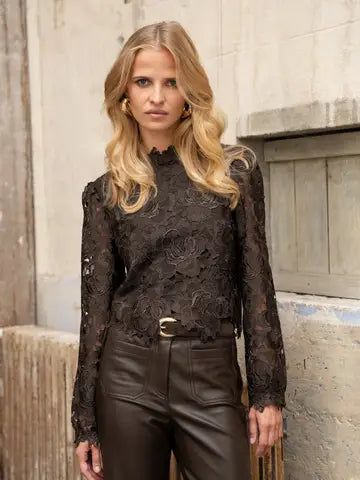 A blonde woman wearing a black lace long-sleeve blouse and dark brown leather pants, standing against a rustic concrete wall.