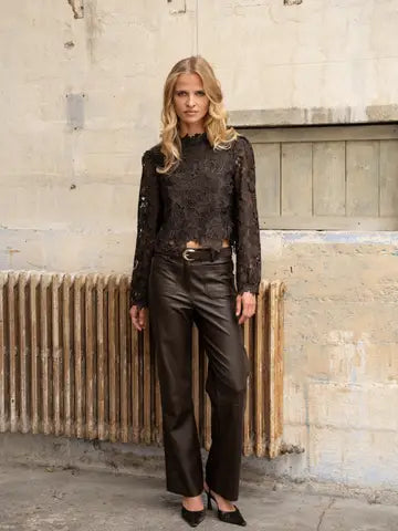 A woman stands outdoors against a weathered beige wall and metal radiator, wearing a black lace long-sleeve top with a high neck, black leather pants, and black pointed shoes.