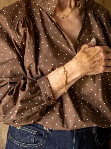 A close-up of a person wearing a brown long-sleeve blouse with a white polka-dot pattern, paired with blue jeans and gold jewelry.