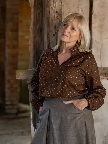 A woman with blonde hair wearing a brown long-sleeve button-up shirt with small white polka dots and a gray skirt, standing in a rustic indoor setting with wooden and stone walls.