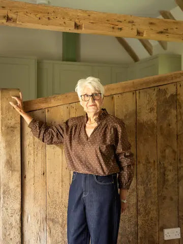 An older woman with short white hair and glasses stands indoors against a rustic wooden wall, wearing a brown patterned blouse and dark blue jeans.