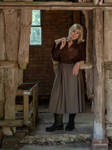 An older woman stands in a rustic wooden doorway of a brick building, wearing a brown patterned blouse, a brown pleated skirt, black boots, and holding a light-colored coat.