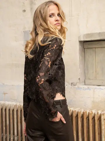 A woman wearing a sheer black lace long-sleeve top and dark brown leather pants, standing in front of a rustic stone wall and radiator.