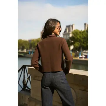 A woman wearing a brown long-sleeve cropped knit top and dark blue jeans, photographed from behind while standing outdoors near a river with a bridge and buildings in the background.