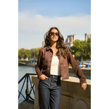 A woman wearing a brown leather jacket, white top, and dark blue jeans stands outdoors by a river with a city skyline in the background.