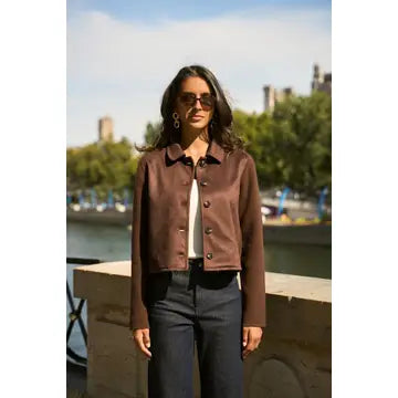 A woman wearing a brown cropped suede jacket, white top, and dark trousers stands outdoors by a river with trees and buildings in the background.