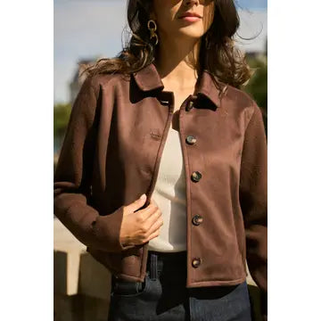 A woman wearing a brown cropped jacket with a collar and dark buttons, layered over a white top and dark jeans, photographed outdoors in natural light.